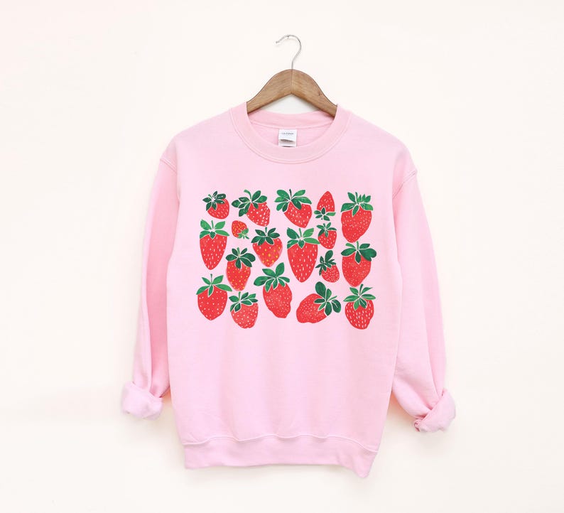 K&ouml;nnte beinhalten: Ein rosa Sweatshirt mit einem grafischen Druck von roten Erdbeeren. Die Erdbeeren sind in einem Muster auf der Vorderseite des Sweatshirts angeordnet.