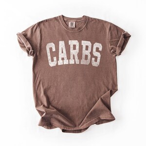 May include: A brown t-shirt with the word "CARBS" printed in large, distressed white letters across the front. The shirt has a crew neck and short sleeves, with the sleeves rolled up. The fabric appears soft and slightly faded.