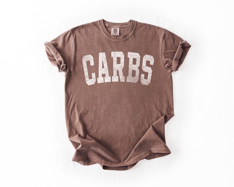 May include: A brown t-shirt with the word "CARBS" printed in large, distressed white letters across the front. The shirt has a crew neck and short sleeves, with the sleeves rolled up. The fabric appears soft and slightly faded.