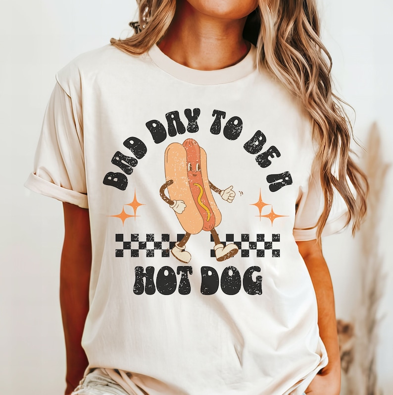 May include: A beige t-shirt with a retro-style graphic of a hot dog with arms and legs. The text "Bad Day To Be A Hot Dog" is printed in black on the shirt.