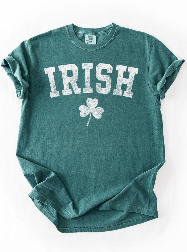 May include: Teal-colored t-shirt with the word "IRISH" in distressed white lettering and a white shamrock graphic. The shirt has a crew neck and short sleeves, perfect for St. Patrick's Day or any day.