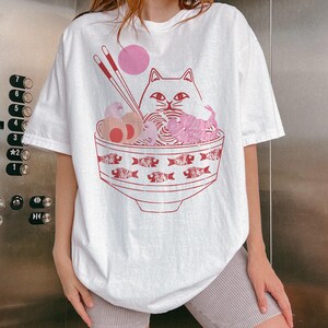 May include: White oversized t-shirt with a graphic of a cat eating ramen noodles from a bowl with fish designs. The cat has pink ears and a pink nose.
