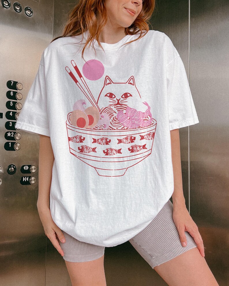 May include: White oversized t-shirt with a graphic of a cat eating ramen noodles from a bowl with fish designs. The cat has pink ears and a pink nose.