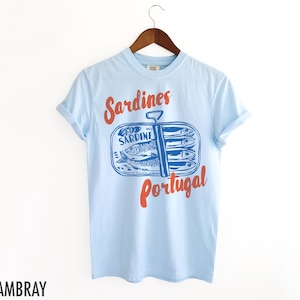 Op de afbeelding: Lichtblauw T-shirt met een afbeelding van een blikje sardines met de tekst "Sardines Portugal" in rood en blauw.