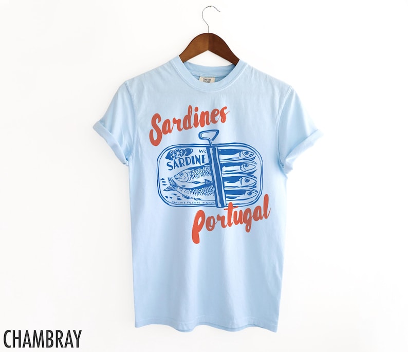 Op de afbeelding: Lichtblauw T-shirt met een afbeelding van een blikje sardines met de tekst "Sardines Portugal" in rood en blauw.