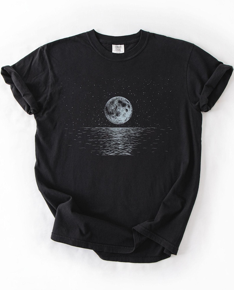 May include: Black t-shirt featuring a detailed illustration of a full moon reflecting on water, set against a starry night sky. The design is in white and gray tones, creating a serene and celestial aesthetic. The shirt has a classic crew neck and short sleeves.