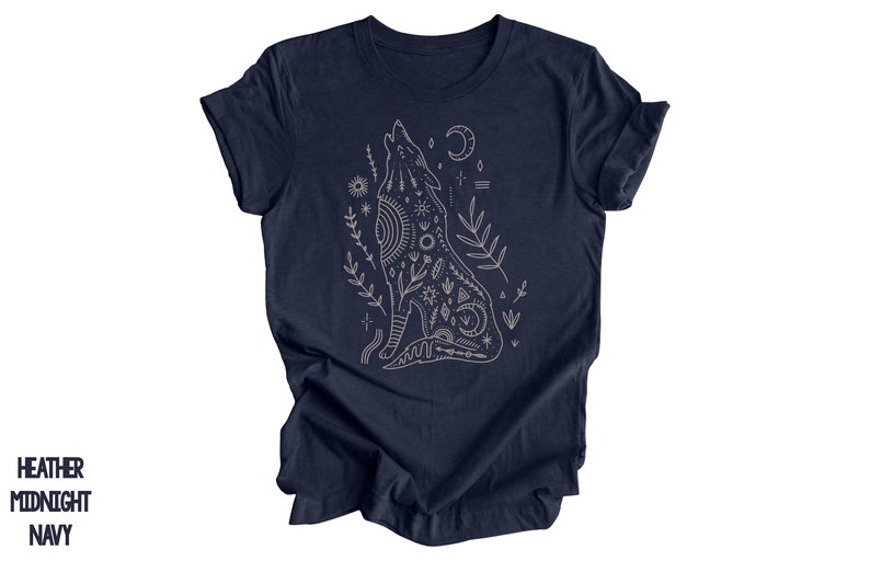 May include: A navy blue t-shirt with a white outline of a howling wolf with intricate geometric patterns and floral accents. The wolf is surrounded by stars, moons, and other celestial symbols.