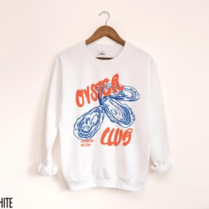 K&ouml;nnte beinhalten: Wei&szlig;er Pullover mit einer blauen und orangenen Grafik von Austern und dem Text "Oyster Club" und "Shucked All Day".