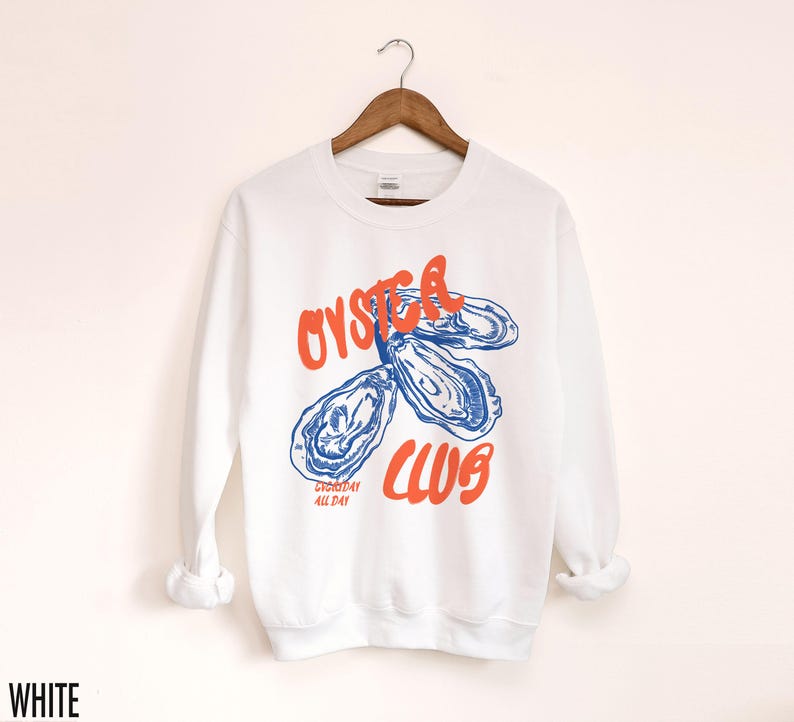 K&ouml;nnte beinhalten: Wei&szlig;er Pullover mit einer blauen und orangenen Grafik von Austern und dem Text "Oyster Club" und "Shucked All Day".
