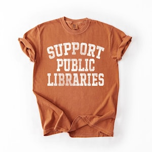 May include: A rust-colored t-shirt with the text "SUPPORT PUBLIC LIBRARIES" in a bold, white, collegiate-style font. The shirt has rolled-up sleeves and is displayed on a white background.