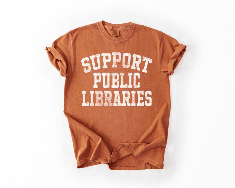 May include: A rust-colored t-shirt with the text "SUPPORT PUBLIC LIBRARIES" in a bold, white, collegiate-style font. The shirt has rolled-up sleeves and is displayed on a white background.