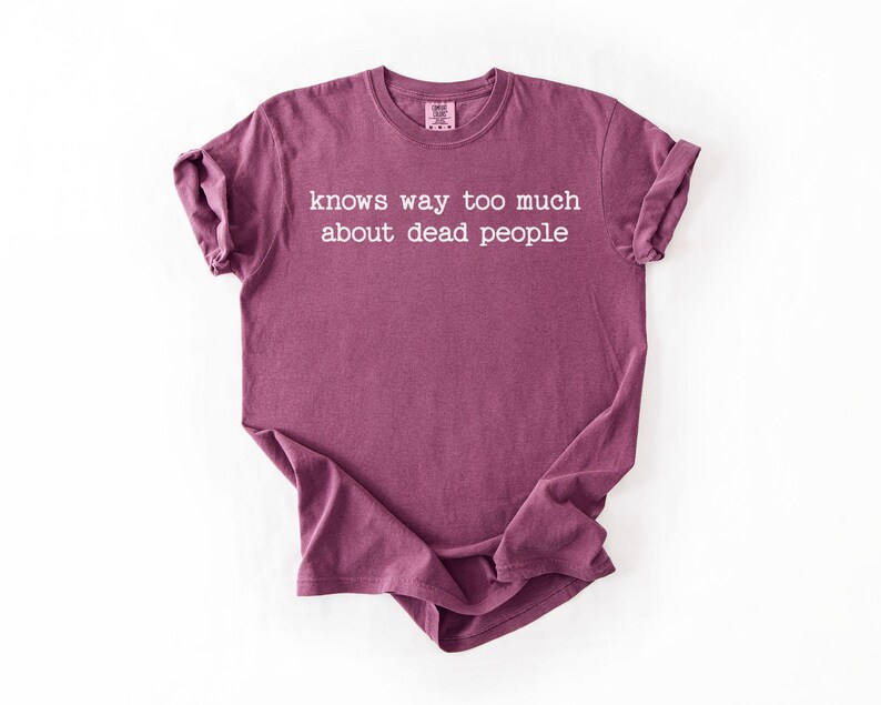 Puede incluir: Una camiseta granate con el texto "knows way too much about dead people" en letras blancas. La camiseta est&aacute; doblada y colocada sobre una superficie blanca. La camiseta tiene mangas cortas y cuello redondo.