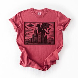 May include: Red t-shirt featuring a black graphic of a dragon perched atop a castle, with a UFO in the sky. The design is a woodcut style illustration. The shirt is made of a soft material.