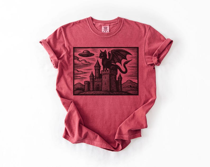 May include: Red t-shirt featuring a black graphic of a dragon perched atop a castle, with a UFO in the sky. The design is a woodcut style illustration. The shirt is made of a soft material.