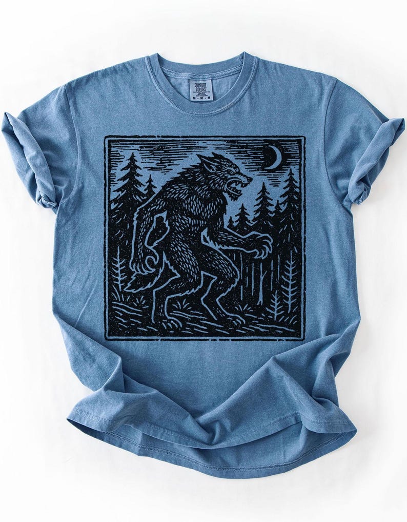 May include: A blue t-shirt featuring a black graphic of a werewolf in a forest under a crescent moon. The werewolf is depicted in a stylized, vintage illustration style. The shirt has a crew neck and short sleeves.