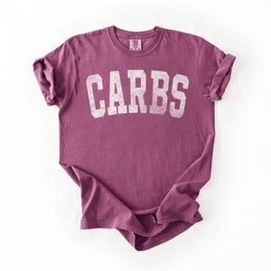 May include: A maroon t-shirt with the word "CARBS" printed in white, block letters. The shirt has short sleeves and a crew neck. The fabric appears soft and slightly faded.