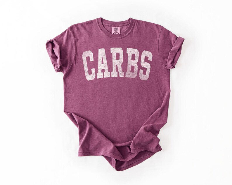 May include: A maroon t-shirt with the word "CARBS" printed in white, block letters. The shirt has short sleeves and a crew neck. The fabric appears soft and slightly faded.