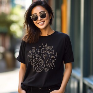 Puede incluir: Una camiseta negra con un dise&ntilde;o gr&aacute;fico dorado que presenta una serpiente, una corona, un diente, un ojo y estrellas.