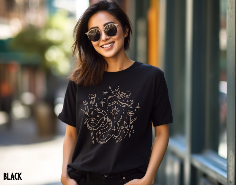 Puede incluir: Una camiseta negra con un dise&ntilde;o gr&aacute;fico dorado que presenta una serpiente, una corona, un diente, un ojo y estrellas.