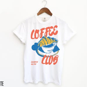 Puede incluir: Camiseta blanca con un gr&aacute;fico de una taza de caf&eacute; con un croissant en la parte superior. El texto "Coffee Club" est&aacute; impreso en rojo y azul. El texto "Everyday All Day" est&aacute; impreso en una fuente m&aacute;s peque&ntilde;a debajo del gr&aacute;fico.