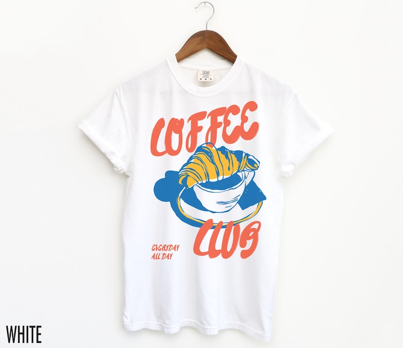 Puede incluir: Camiseta blanca con un gr&aacute;fico de una taza de caf&eacute; con un croissant en la parte superior. El texto "Coffee Club" est&aacute; impreso en rojo y azul. El texto "Everyday All Day" est&aacute; impreso en una fuente m&aacute;s peque&ntilde;a debajo del gr&aacute;fico.