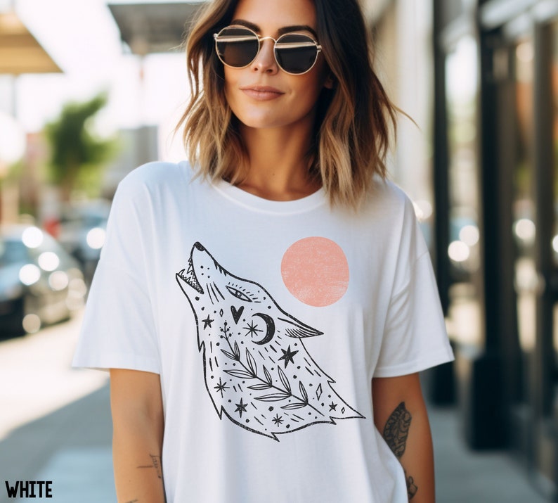 May include: White t-shirt featuring a black and white illustration of a wolf howling at the moon. The wolf is surrounded by stars, leaves, and a crescent moon. The shirt also features a pink circle.