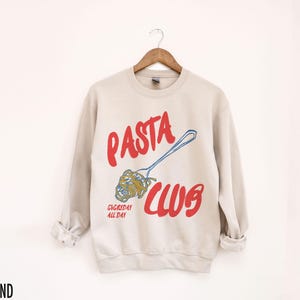May include: A beige sweatshirt with the text "PASTA CLUB" in red and blue. The text "EVERYDAY ALL DAY" is printed in red below the main text. A cartoon image of a fork with spaghetti on it is printed next to the text.