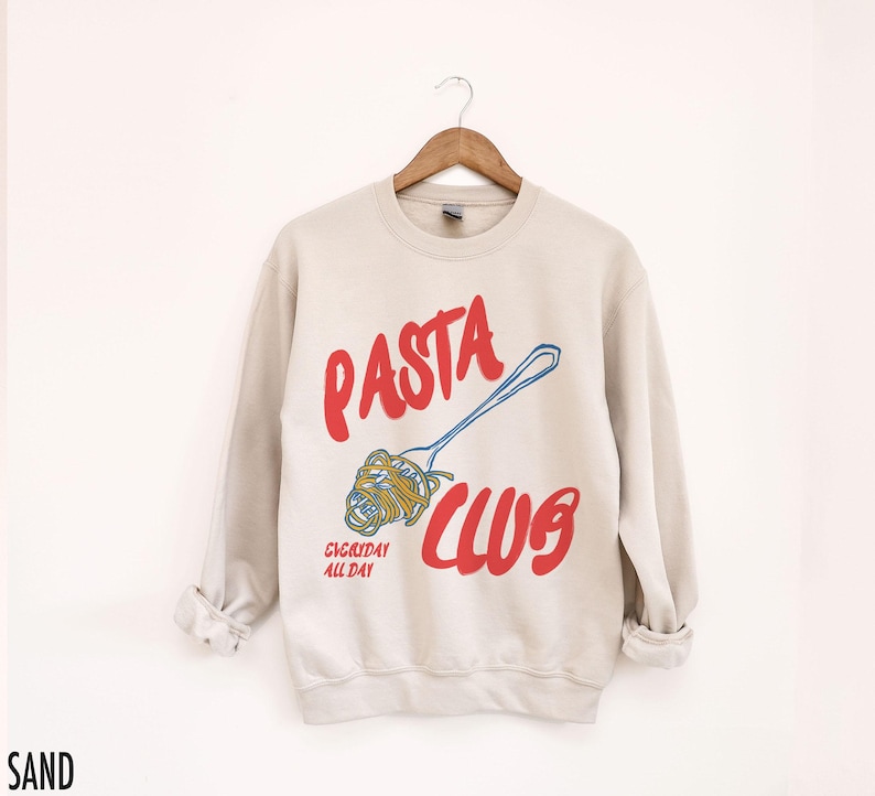 May include: A beige sweatshirt with the text "PASTA CLUB" in red and blue. The text "EVERYDAY ALL DAY" is printed in red below the main text. A cartoon image of a fork with spaghetti on it is printed next to the text.