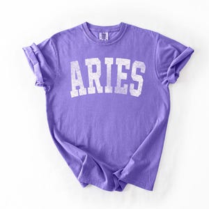 May include: A lavender t-shirt with the word "ARIES" printed in distressed white block letters. The shirt is made of soft material and has rolled-up sleeves. The t-shirt is laid flat on a white surface.