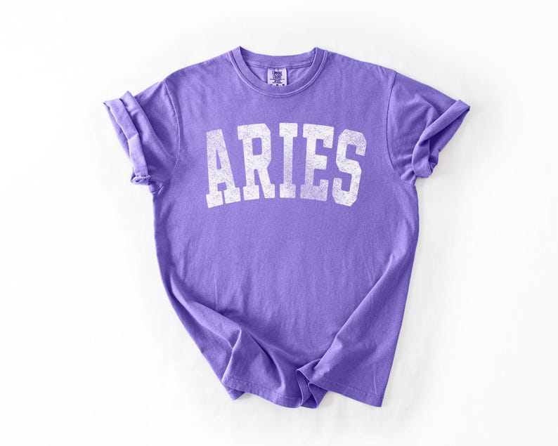 May include: A lavender t-shirt with the word "ARIES" printed in distressed white block letters. The shirt is made of soft material and has rolled-up sleeves. The t-shirt is laid flat on a white surface.