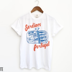 Op de afbeelding: Wit T-shirt met een blauwe en rode afbeelding van een sardineblik met de tekst "Sardines Portugal".