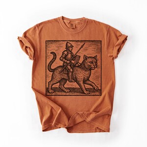 Puede incluir: Una camiseta de color naranja quemado con una ilustraci&oacute;n en blanco y negro de un caballero montando un gato grande. El caballero sostiene una lanza y un escudo. El gato tiene un collar. La camiseta tiene cuello redondo y mangas enrolladas.