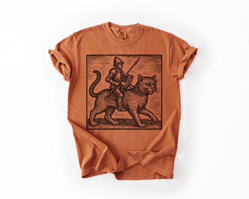 Puede incluir: Una camiseta de color naranja quemado con una ilustraci&oacute;n en blanco y negro de un caballero montando un gato grande. El caballero sostiene una lanza y un escudo. El gato tiene un collar. La camiseta tiene cuello redondo y mangas enrolladas.