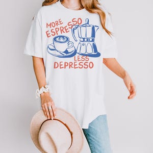 K&ouml;nnte beinhalten: Wei&szlig;es T-Shirt mit einem Kaffee-Motiv. Das Design zeigt eine Kaffeetasse, einen Espressokocher und den Text "MORE ESPRESSO LESS DEPRESSO" in Rot und Blau. Das Shirt wird mit einer blauen Jeans und einem beigen Hut getragen.