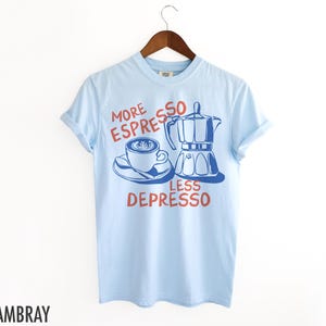 May include: Light blue t-shirt with a graphic of a coffee cup and a coffee pot. The text on the shirt reads "More Espresso Less Depresso".