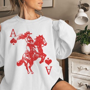 May include: White sweatshirt featuring a red graphic of a woman riding a horse, with the Ace of Spades and Ace of Hearts playing card symbols. The word "WHITE" is printed at the bottom right.