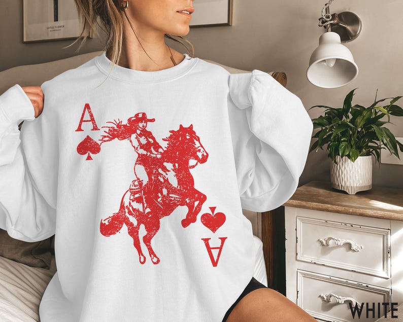 May include: White sweatshirt featuring a red graphic of a woman riding a horse, with the Ace of Spades and Ace of Hearts playing card symbols. The word "WHITE" is printed at the bottom right.