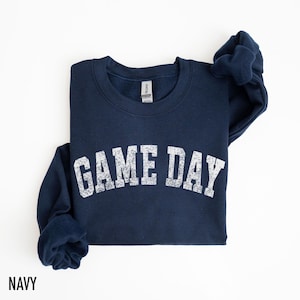 May include: Navy blue sweatshirt with the text "GAME DAY" in distressed white lettering. The sweatshirt is a crew neck style and is folded on a white surface. The word "NAVY" is printed at the bottom left.