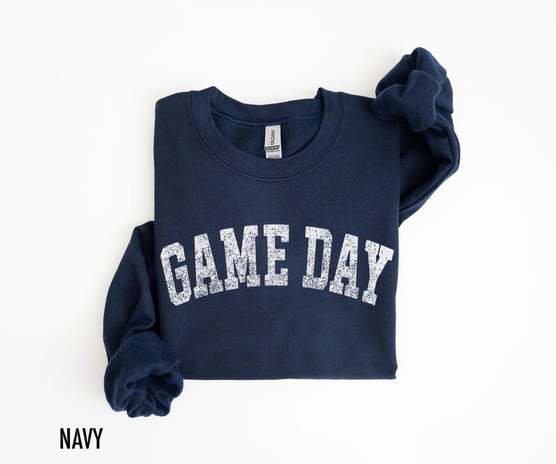 May include: Navy blue sweatshirt with the text "GAME DAY" in distressed white lettering. The sweatshirt is a crew neck style and is folded on a white surface. The word "NAVY" is printed at the bottom left.