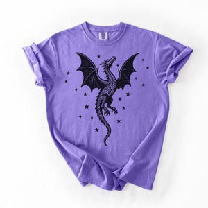 Op de afbeelding: Een lila T-shirt met een zwart drakenontwerp. De draak heeft grote vleugels en is omringd door sterren. Het T-shirt heeft een ronde hals en opgerolde mouwen. Het ontwerp is gecentreerd.