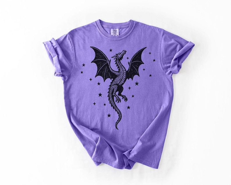 Op de afbeelding: Een lila T-shirt met een zwart drakenontwerp. De draak heeft grote vleugels en is omringd door sterren. Het T-shirt heeft een ronde hals en opgerolde mouwen. Het ontwerp is gecentreerd.
