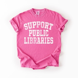 May include: A bright pink t-shirt with the words "SUPPORT PUBLIC LIBRARIES" printed in large, distressed white letters. The shirt has short sleeves and a crew neck. The fabric appears soft and comfortable.