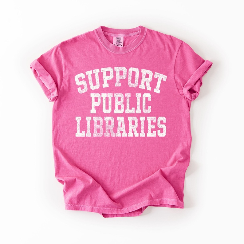 May include: A bright pink t-shirt with the words "SUPPORT PUBLIC LIBRARIES" printed in large, distressed white letters. The shirt has short sleeves and a crew neck. The fabric appears soft and comfortable.