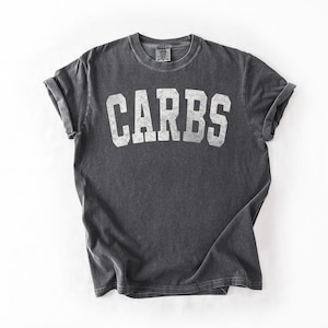 May include: Dark grey t-shirt with rolled sleeves. The word "CARBS" is printed in large, white, block letters across the front. The shirt is made of a soft, comfortable material.