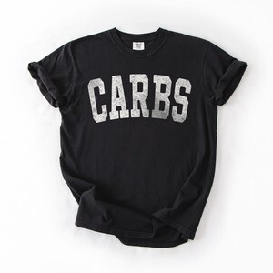 May include: Black t-shirt with the word "CARBS" printed in distressed white block letters. The shirt has short sleeves and a crew neck. The t-shirt is made of a soft material.
