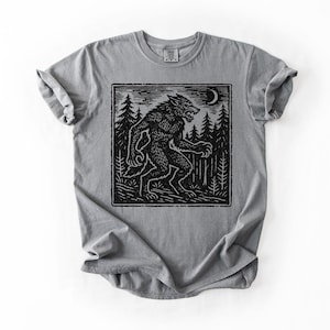 Puede incluir: Camiseta gris con un dise&ntilde;o gr&aacute;fico negro de un hombre lobo en un bosque bajo una luna creciente. El dise&ntilde;o es de estilo xilograf&iacute;a. Cuello redondo y mangas cortas.