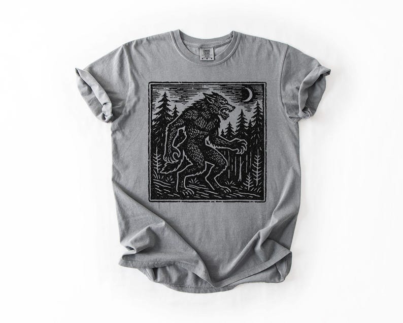 Puede incluir: Camiseta gris con un dise&ntilde;o gr&aacute;fico negro de un hombre lobo en un bosque bajo una luna creciente. El dise&ntilde;o es de estilo xilograf&iacute;a. Cuello redondo y mangas cortas.