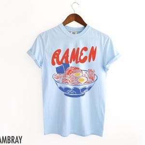 May include: Light blue t-shirt with a red and blue illustration of a bowl of ramen noodles. The text "RAMEN" is printed in red above the bowl.