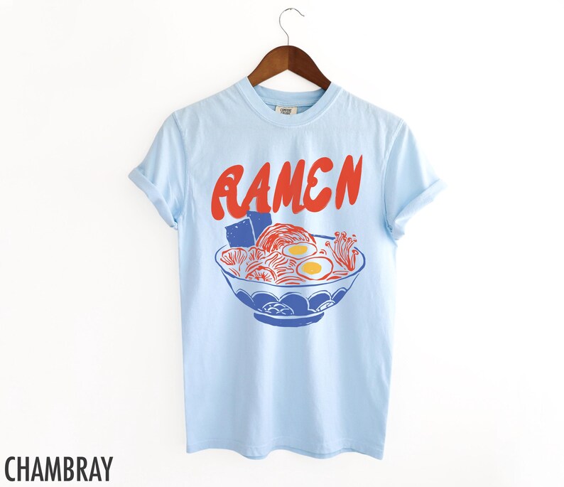 May include: Light blue t-shirt with a red and blue illustration of a bowl of ramen noodles. The text "RAMEN" is printed in red above the bowl.