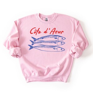 K&ouml;nnte beinhalten: Ein hellrosa Sweatshirt mit hochgekrempelten &Auml;rmeln. Auf der Vorderseite steht der Text "C&ocirc;te d'Azur" in Rot &uuml;ber drei blauen Fischillustrationen. Es ist ein l&auml;ssiges Kleidungsst&uuml;ck.